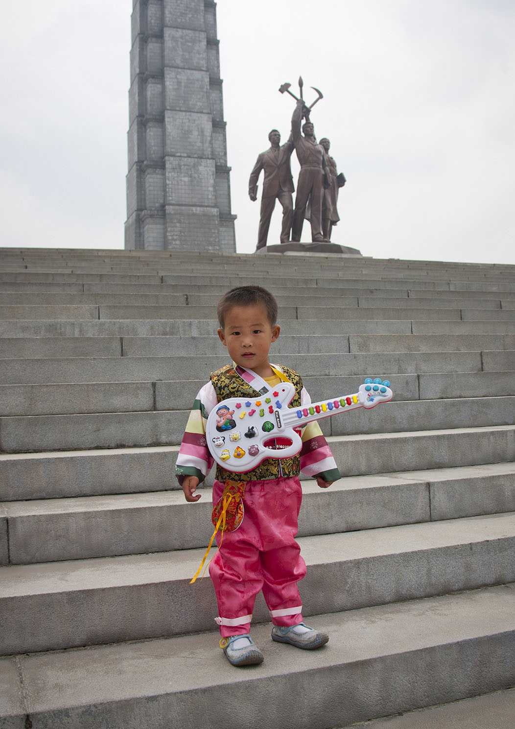 Young Boy With Toy Guitar On Stairs In Front Of Juche Tower, Pyongyang, North Korea