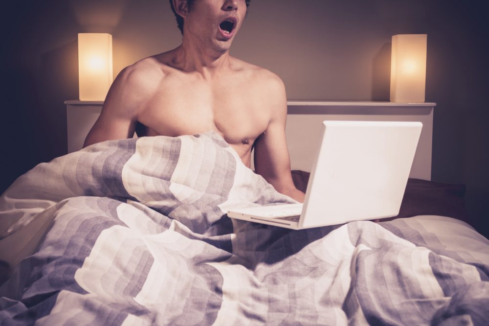 Young man is sitting in bed and watching pornography on laptop