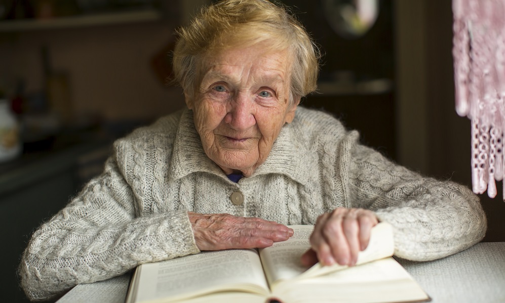 Old woman reading a book sitting at the table.